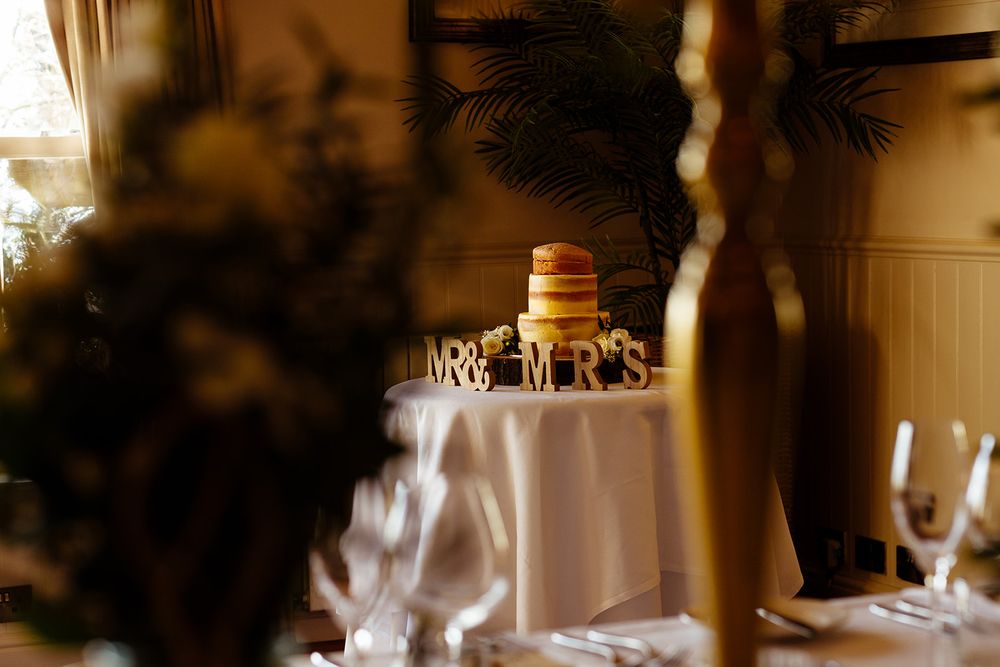 Wedding cake with Mr & Mrs topper on a white draped table, seen through elegant glassware in a warmly lit reception venue.