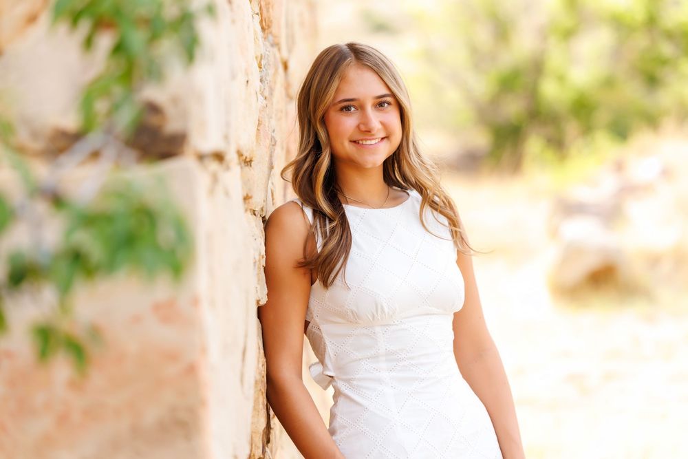 Young woman in white dress smiling against stone wall outdoors in warm sunlight.