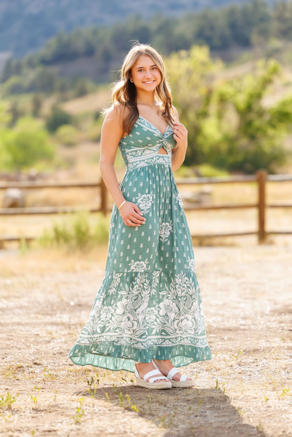 Young woman in a green floral maxi dress and white sandals posing outdoors with mountains in the background.