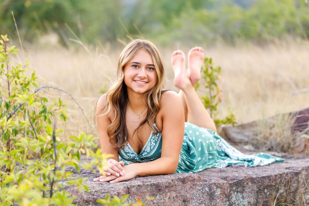 Young woman in teal floral dress lying on rock wall, smiling outdoors in natural field setting.