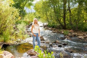 Blonde woman in white top and jeans standing on rocks by a flowing mountain stream surrounded by lush green trees.