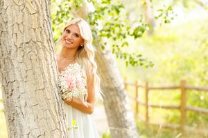 Smiling blonde woman in a mint dress holding a bouquet, posing beside a tree in a sunny outdoor setting.
