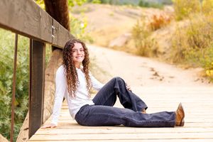 Young woman with curly hair sitting on a wooden boardwalk, smiling outdoors in jeans and white shirt.