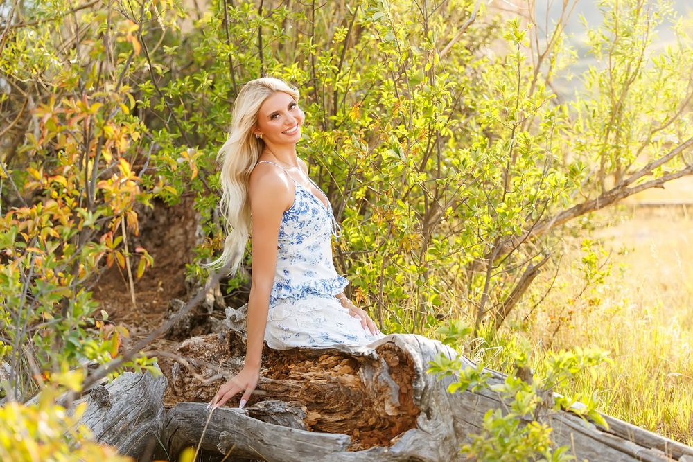 Smiling blonde woman in floral dress sitting on a log surrounded by lush green and yellow foliage outdoors.