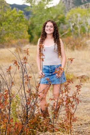 A smiling teenage girl with curly hair wearing a striped tank top and denim skirt stands outdoors among autumn foliage.