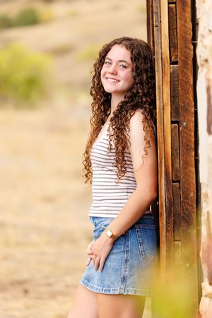 Smiling teenage girl with curly hair in striped top and denim skirt leaning against rustic wooden door outdoors.