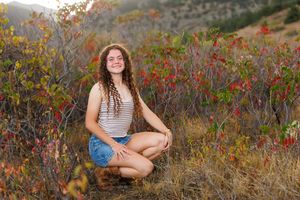 Young woman with curly hair smiling in a field of colorful autumn foliage, wearing denim shorts and striped top.