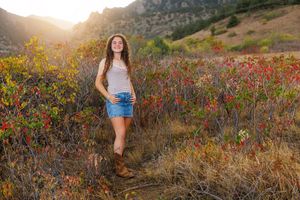 Young woman in denim shorts and boots smiling in autumn mountain landscape with colorful foliage at sunset.