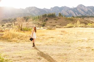 Young woman in white dress walking through golden meadow with Flatirons mountain range in background.