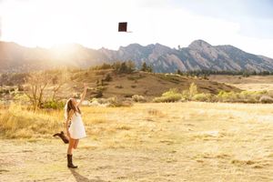 A young woman in a white dress and cowboy boots tosses her graduation cap in a sunlit mountain meadow.