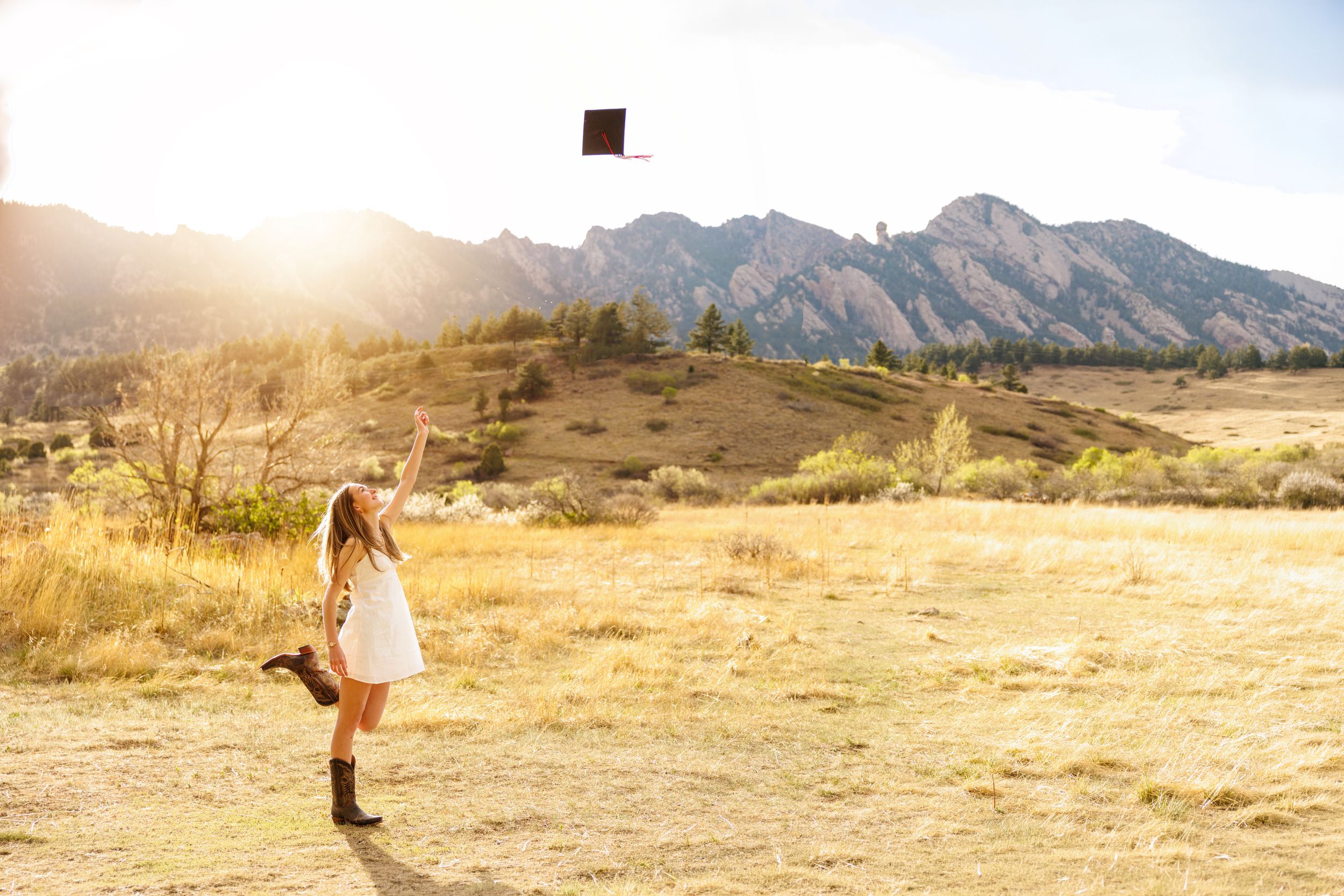 A young woman in a white dress and cowboy boots tosses her graduation cap in a sunlit mountain meadow.