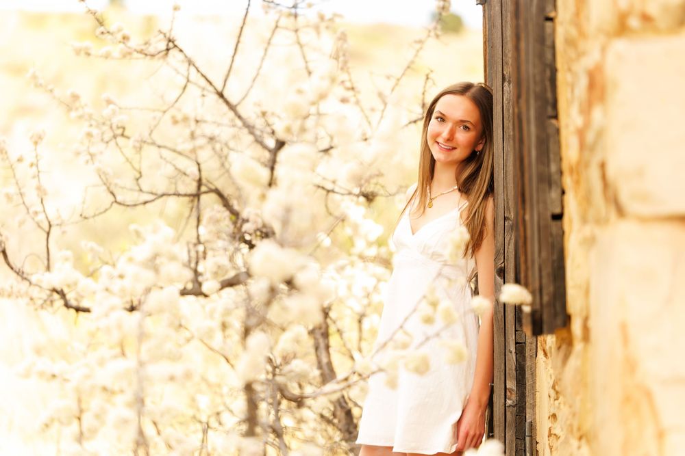 Young woman in white dress smiling outdoors near blooming white spring trees in warm sunlight.