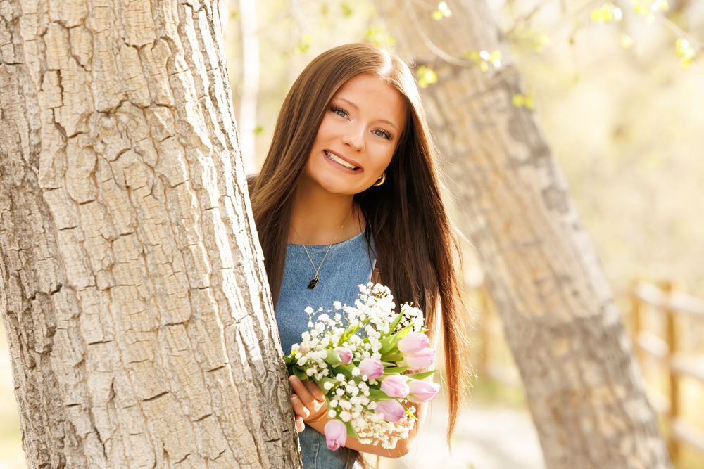 Young girl with long brown hair smiling behind a tree, holding a bouquet of white and pink flowers outdoors.