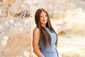 Young woman with long brown hair in a blue dress poses outdoors near a stone wall with white blossoms.