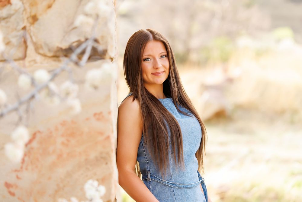 Young woman with long brown hair in a blue dress poses outdoors near a stone wall with white blossoms.