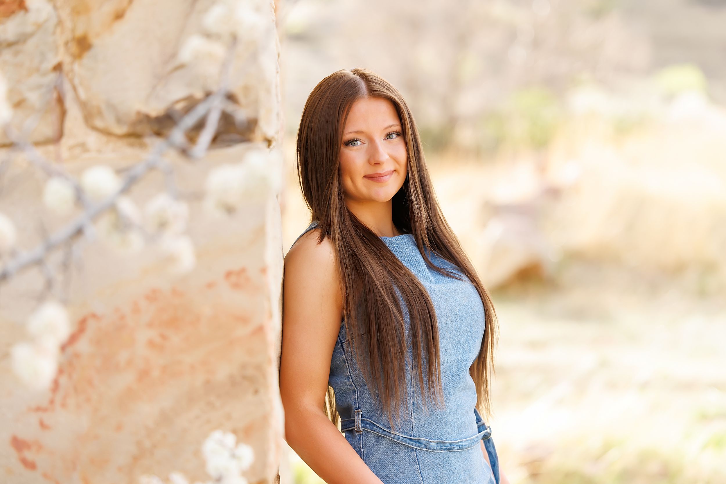 Young woman with long brown hair in a blue dress poses outdoors near a stone wall with white blossoms.