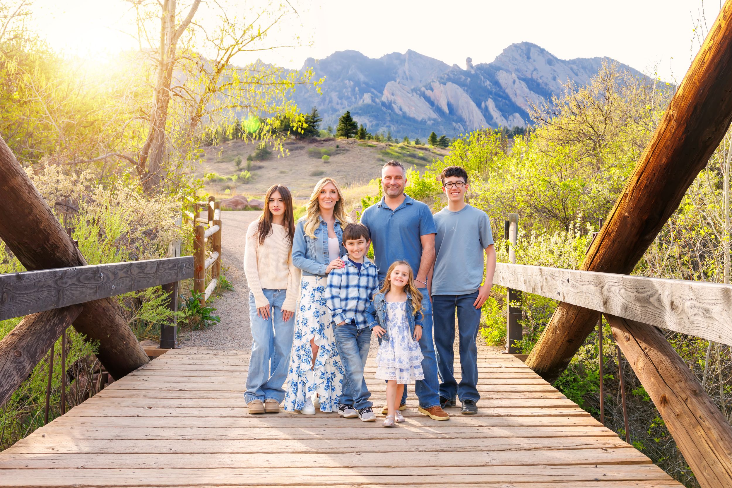 Family of six poses on a wooden bridge with Flatirons mountains and golden sunset in Boulder, Colorado.