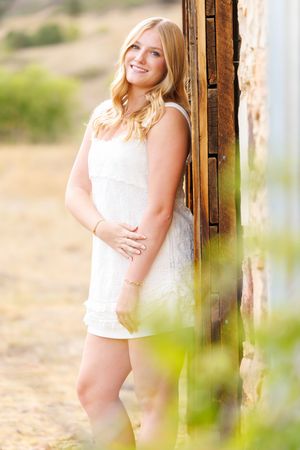 Smiling blonde woman in white lace dress leaning against rustic wooden door in sunny outdoor setting.