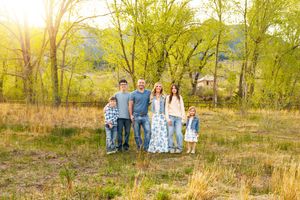 Family of six posing outdoors in a sunny meadow with lush green trees in the background during spring.
