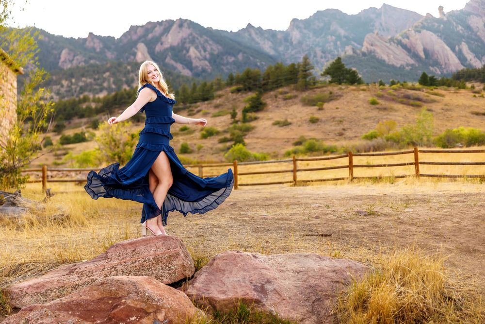 Woman in flowing blue dress dancing on rocks with Rocky Mountain flatirons and wooden fence in background.