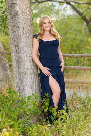 Young blonde woman in navy ruffled maxi dress poses by a tree in a lush outdoor setting.
