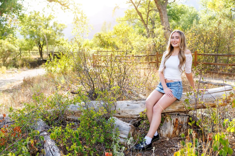 Young woman in white top and denim shorts sitting on a log in a sunny, lush green outdoor nature setting.