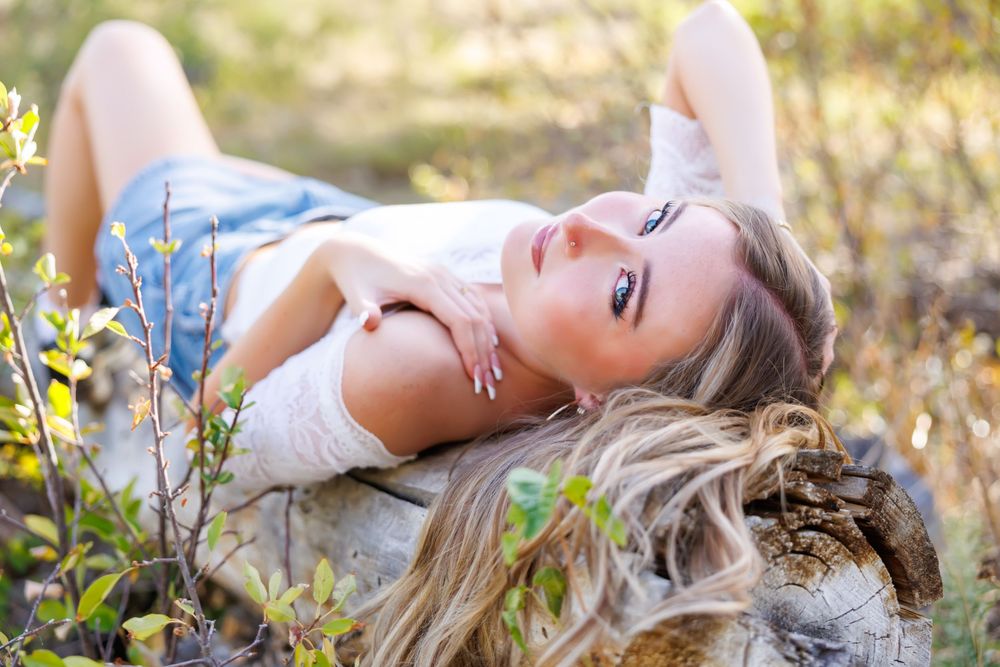 Young blonde woman lying on the ground outdoors among green plants in warm sunlight, wearing casual denim shorts.