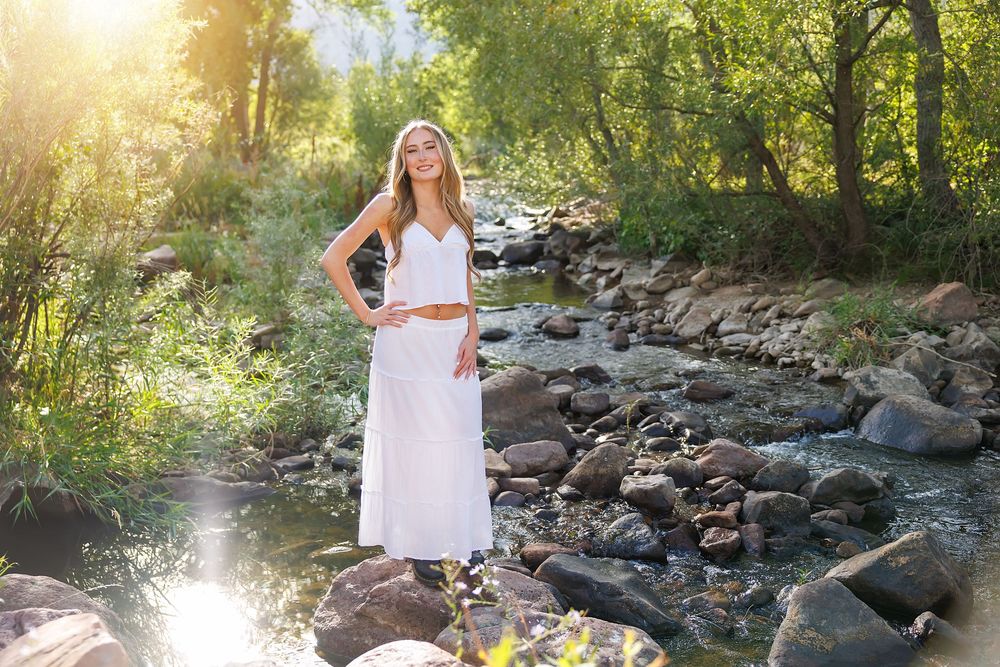 A young woman in a white maxi dress stands gracefully on rocks beside a sunlit forest stream.