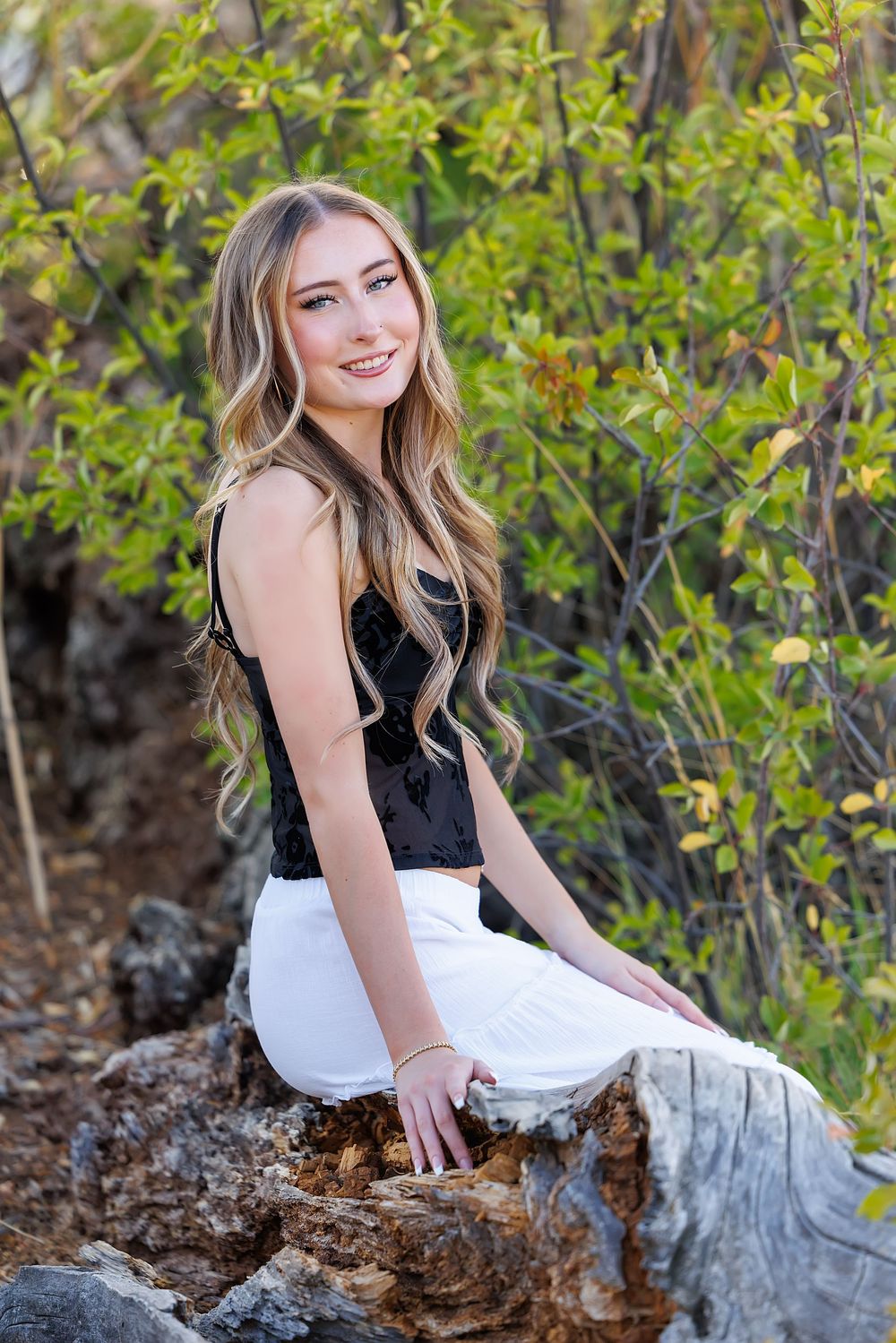 Young blonde woman in black top and white pants sitting on a rock surrounded by green foliage outdoors.