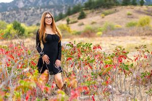 Young woman in black dress and glasses poses among vibrant red autumn shrubs with mountain backdrop.