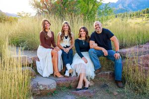 A group sits casually along a stone wall with tall grasses and mountains in the distance against a bright blue sky.