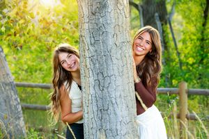 Two friends peek playfully from behind a large tree trunk in a sunny outdoor setting with green foliage.
