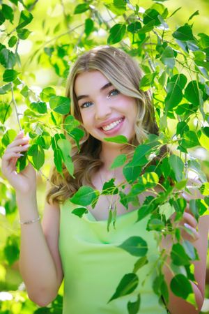 A woman smiles through green leaves in bright natural lighting.