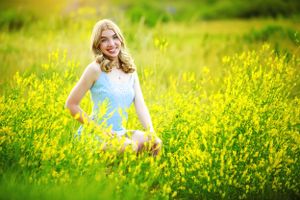 A woman in a blue dress poses in a field of bright yellow wildflowers on a sunny day.