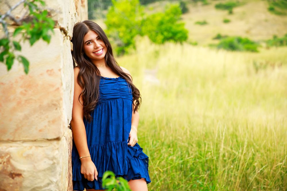 A person in a blue dress poses outdoors in a sunny field with natural grass and trees in the background.