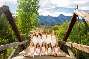 Group in white dresses posed on wooden bridge with mountain scenery.