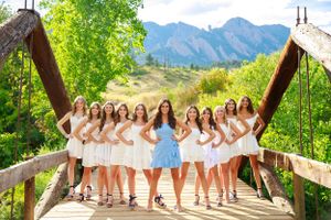 A group in white dresses poses on a wooden bridge with mountain scenery in the background.