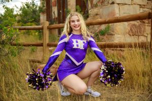 A cheerleader in purple uniform poses with pom poms outdoors.