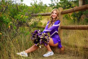 A cheerleader poses with purple pom poms near a wooden fence in a field.