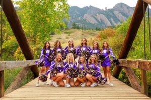 A cheerleading squad in purple uniforms poses on a wooden bridge with mountains behind them.