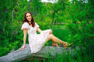 A dreamy summer portrait shows someone in a flowy white dress posing on a fallen log amid verdant foliage.