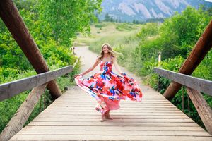A flowing floral red and blue dress twirls on a wooden footbridge with mountain scenery and green trees in the background.