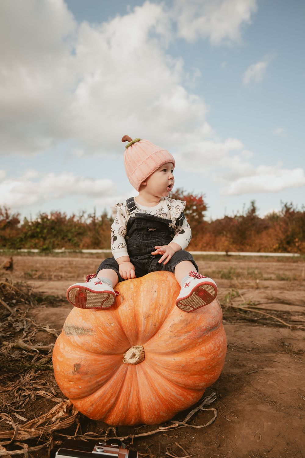 Baby in pink beanie sitting on giant orange pumpkin against cloudy sky.