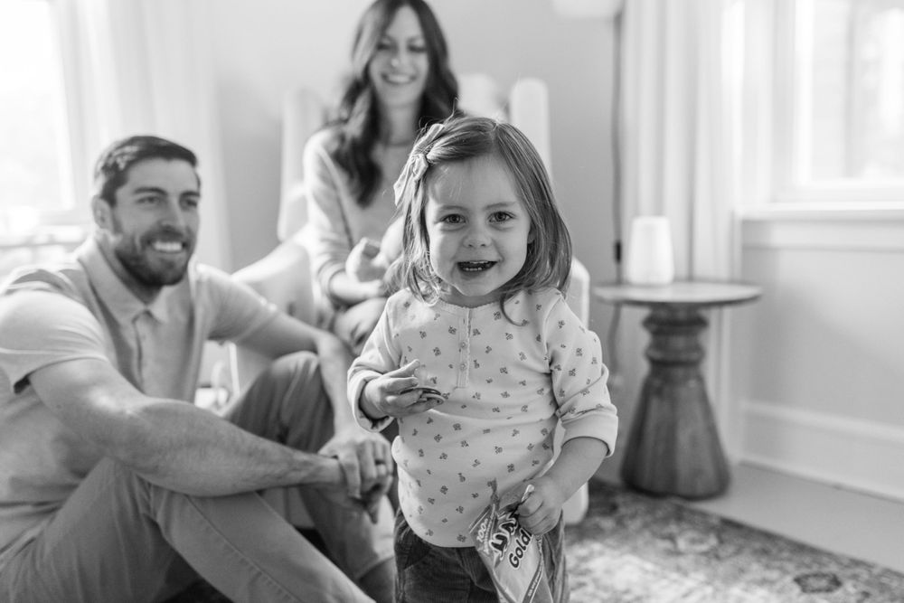 Black and white photo of happy toddler girl smiling at camera with parents sitting behind her.