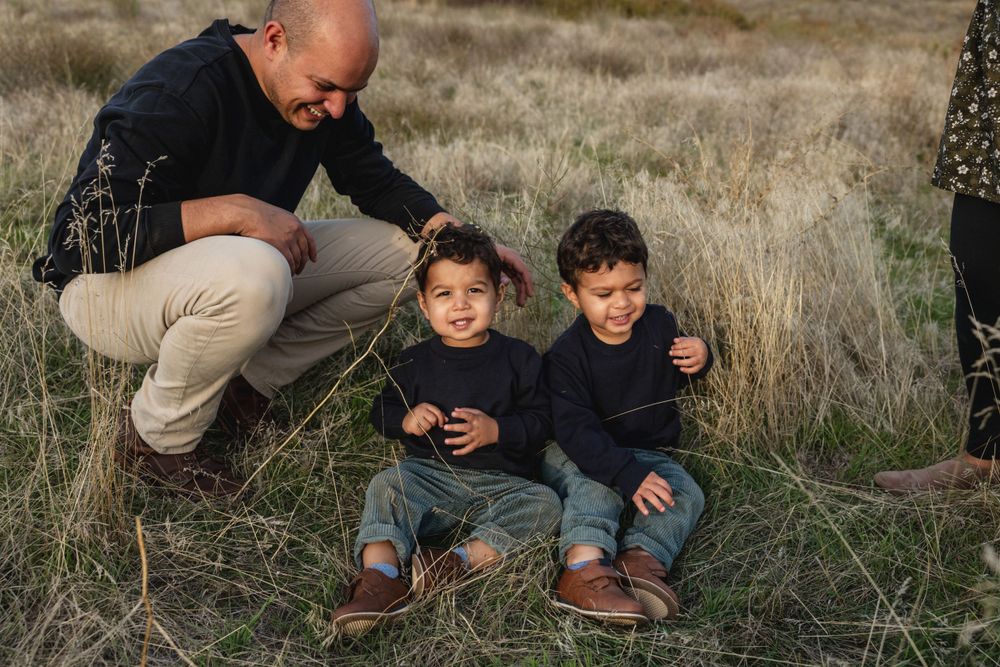 Father crouching with two smiling young boys sitting in dry grass in a field.