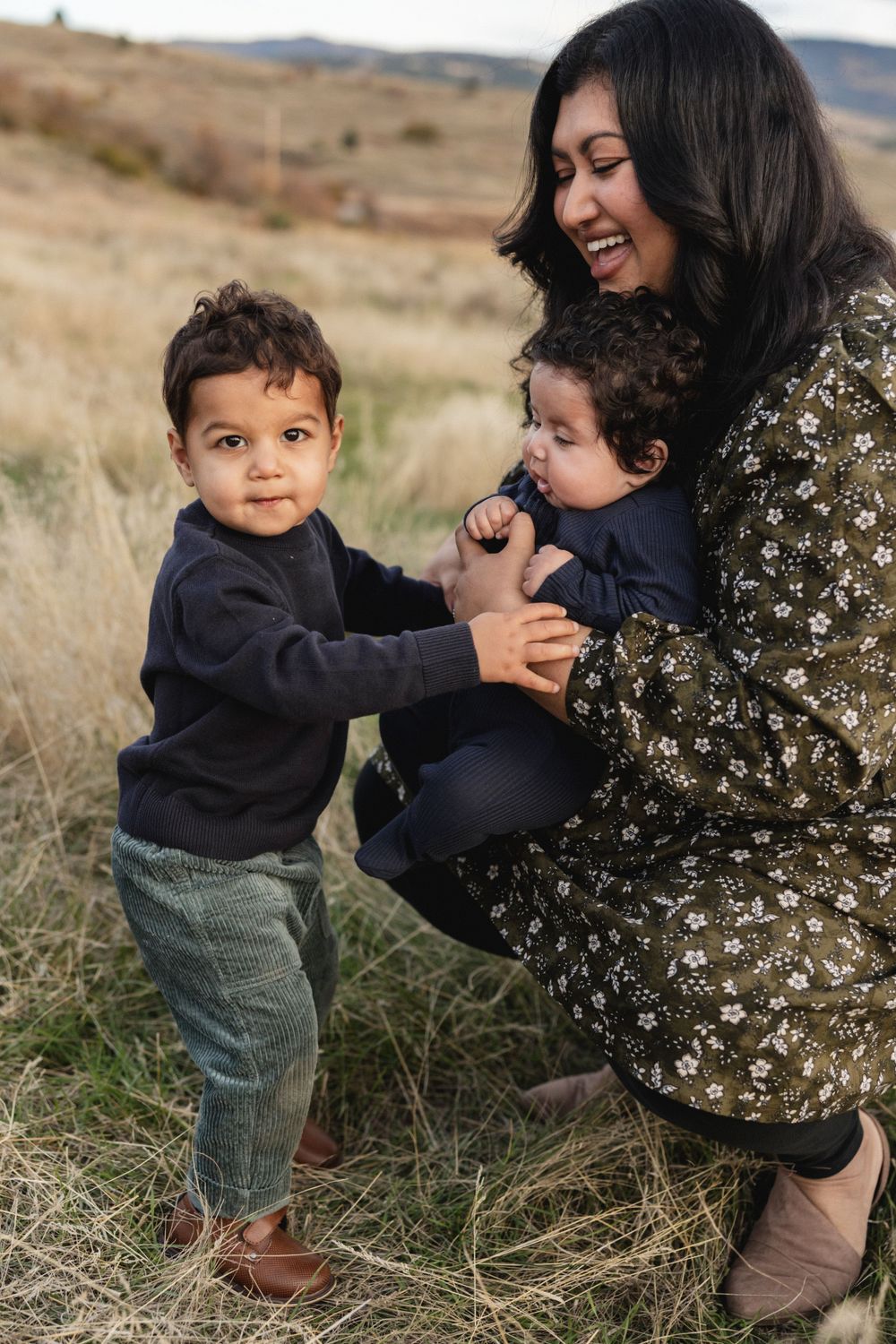 Laughing mother in floral dress crouching with two young sons in a grassy field.