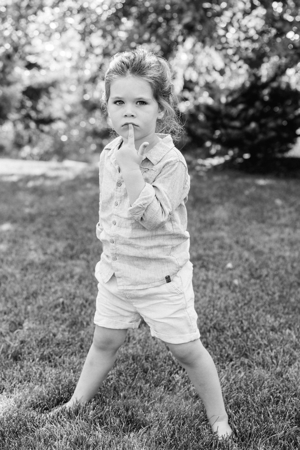 Black and white photo of a young child standing barefoot on grass in a garden, wearing shorts and a button-up shirt.