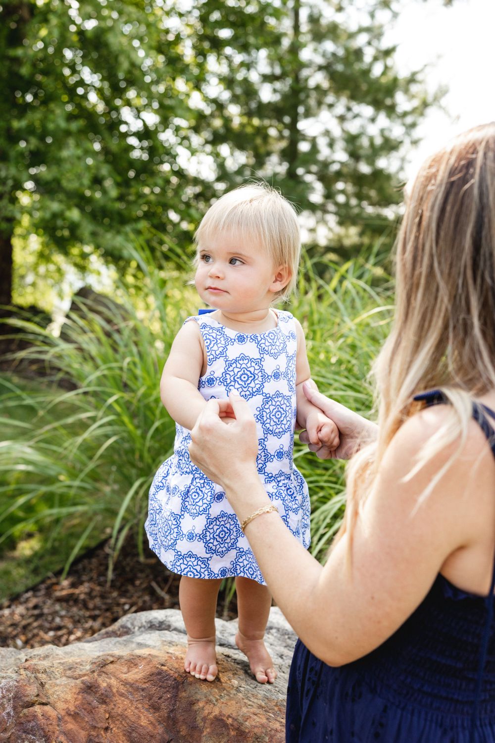 A mother holds her baby girl in a blue patterned dress while standing on rocks surrounded by lush greenery.
