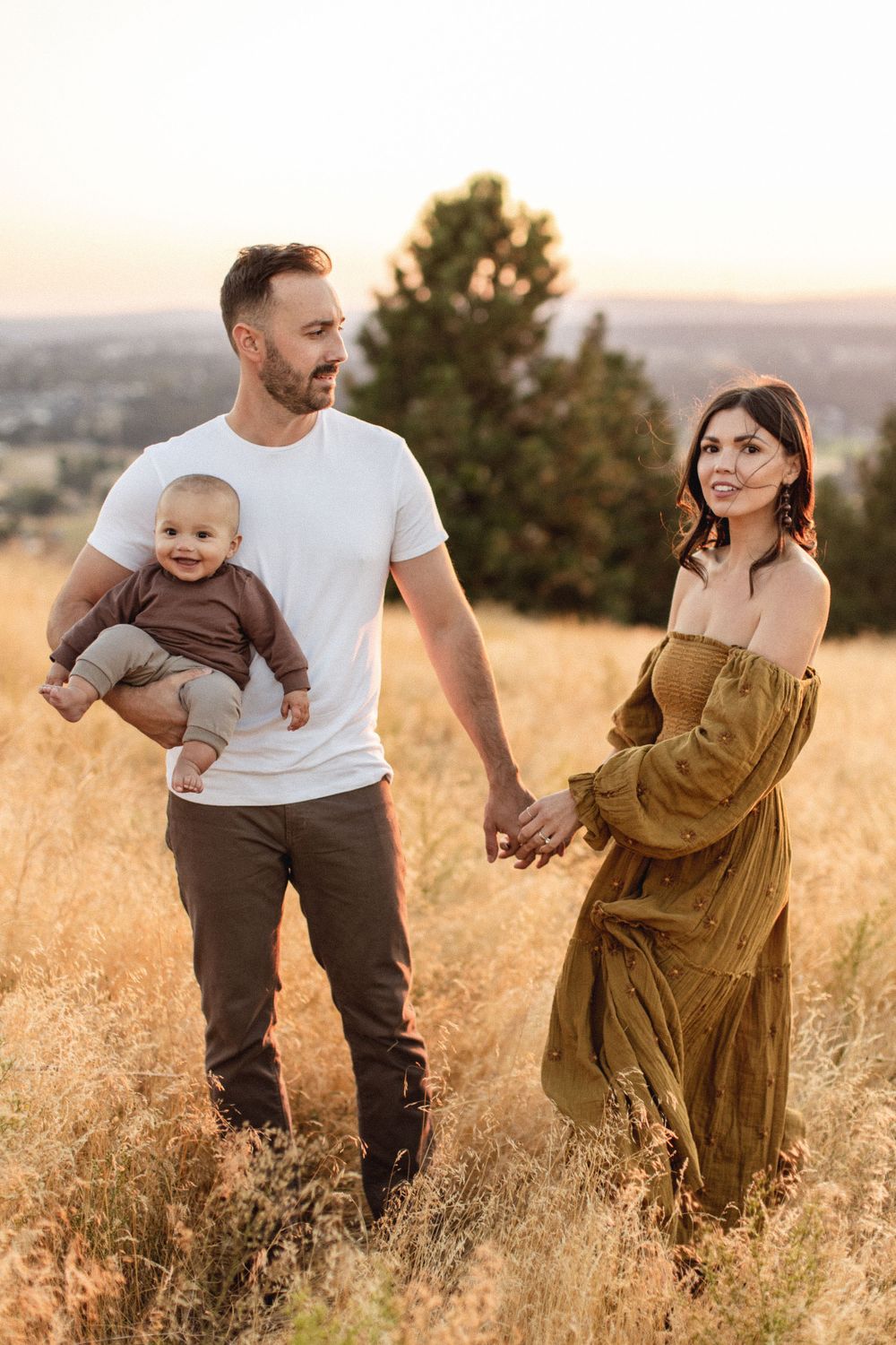 A couple holding hands and carrying their baby walks through a golden field at sunset.