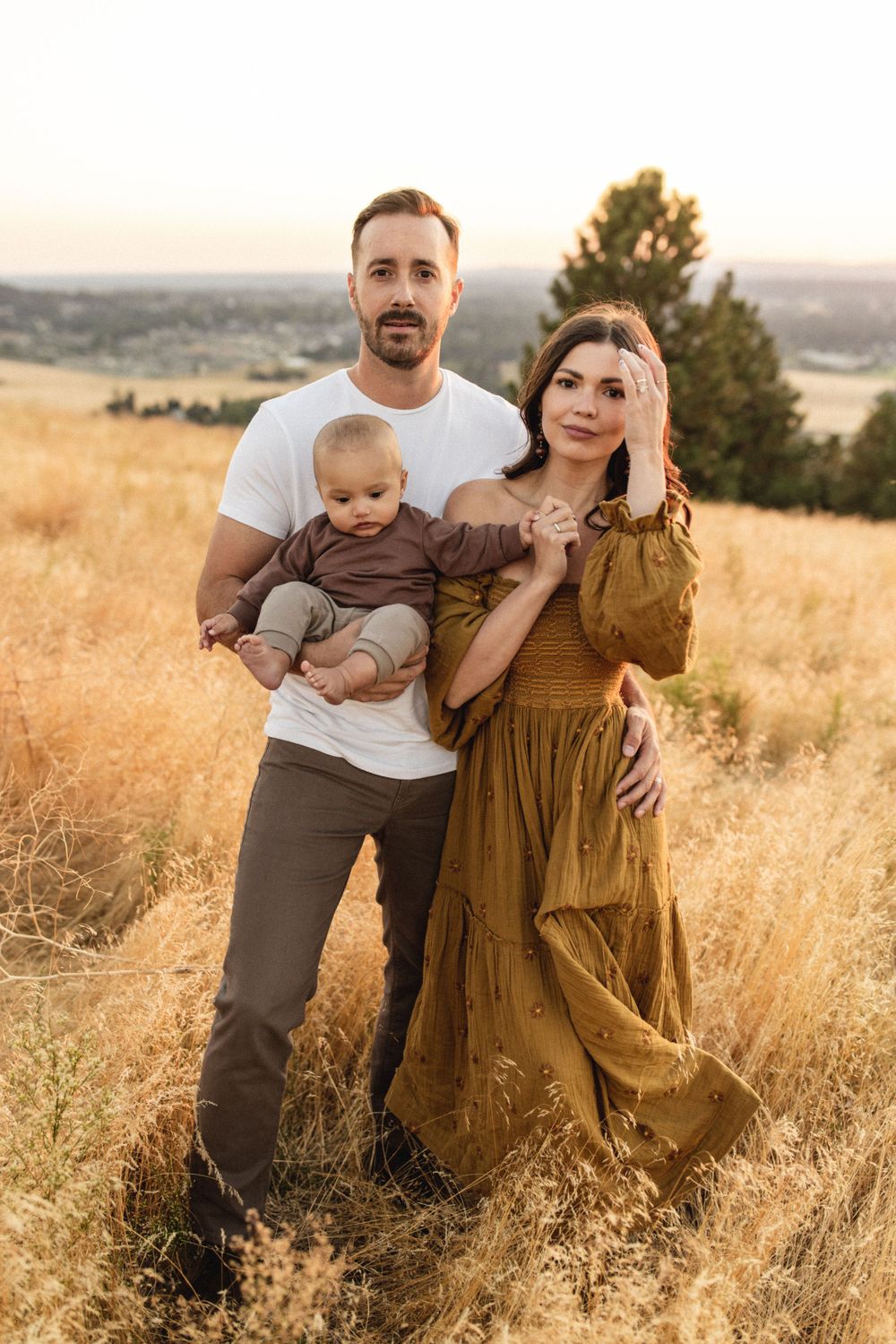 A family of three poses in a golden meadow at sunset, father holding baby in a brown outfit.